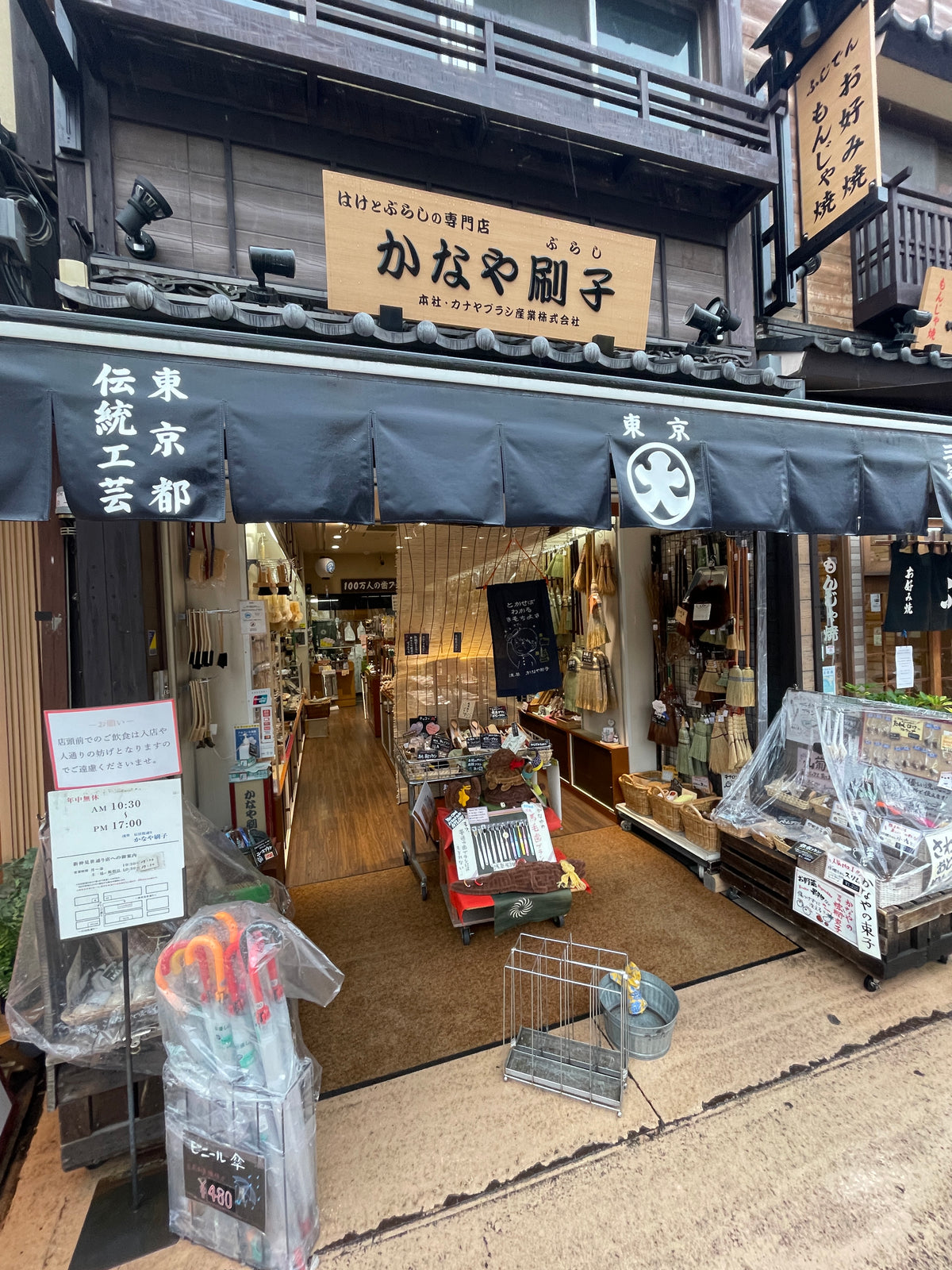 Street view of a traditional Japanese shop with black awnings and wooden signage, featuring the Japanese Mystery Box—an assortment of unique gifts and souvenirs displayed invitingly at the entrance and inside the well-lit store.