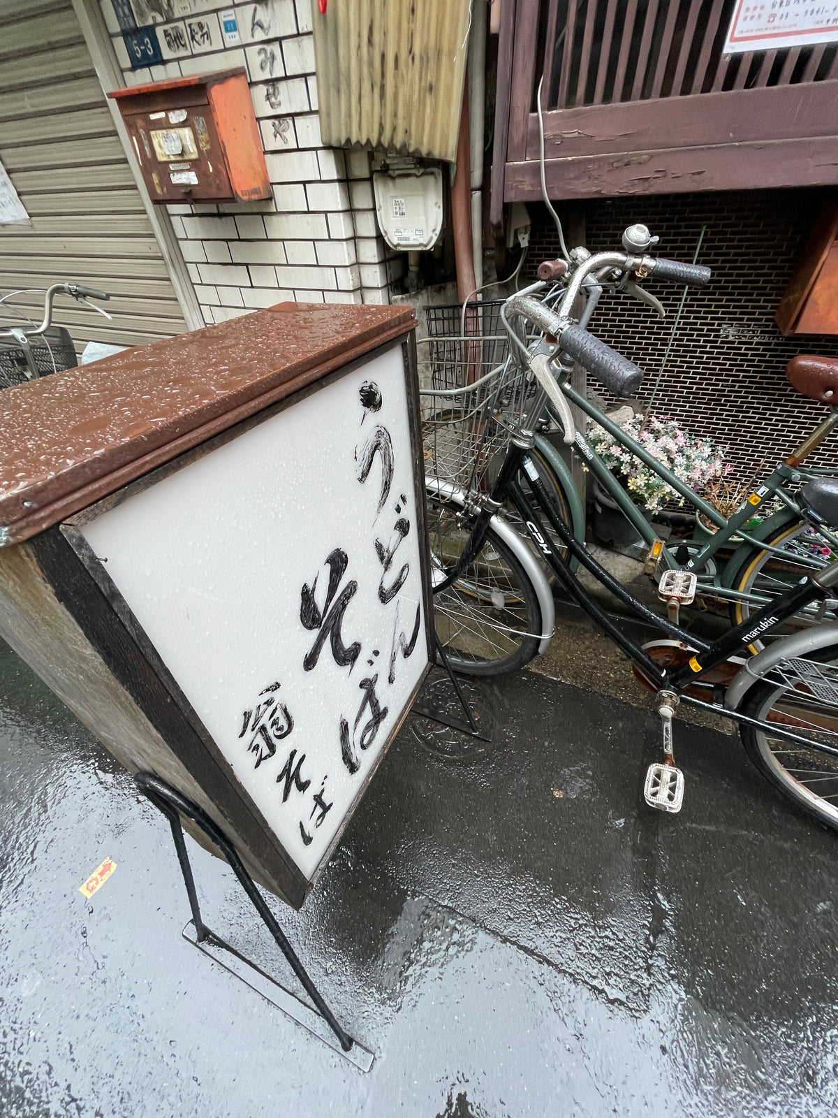 A sign with Japanese text stands on a wet sidewalk next to parked bicycles on a rainy day. It reads うどん そば (udon, soba), inviting travelers to discover the Japanese Mystery Box for unique gifts and travel souvenirs from Japan.