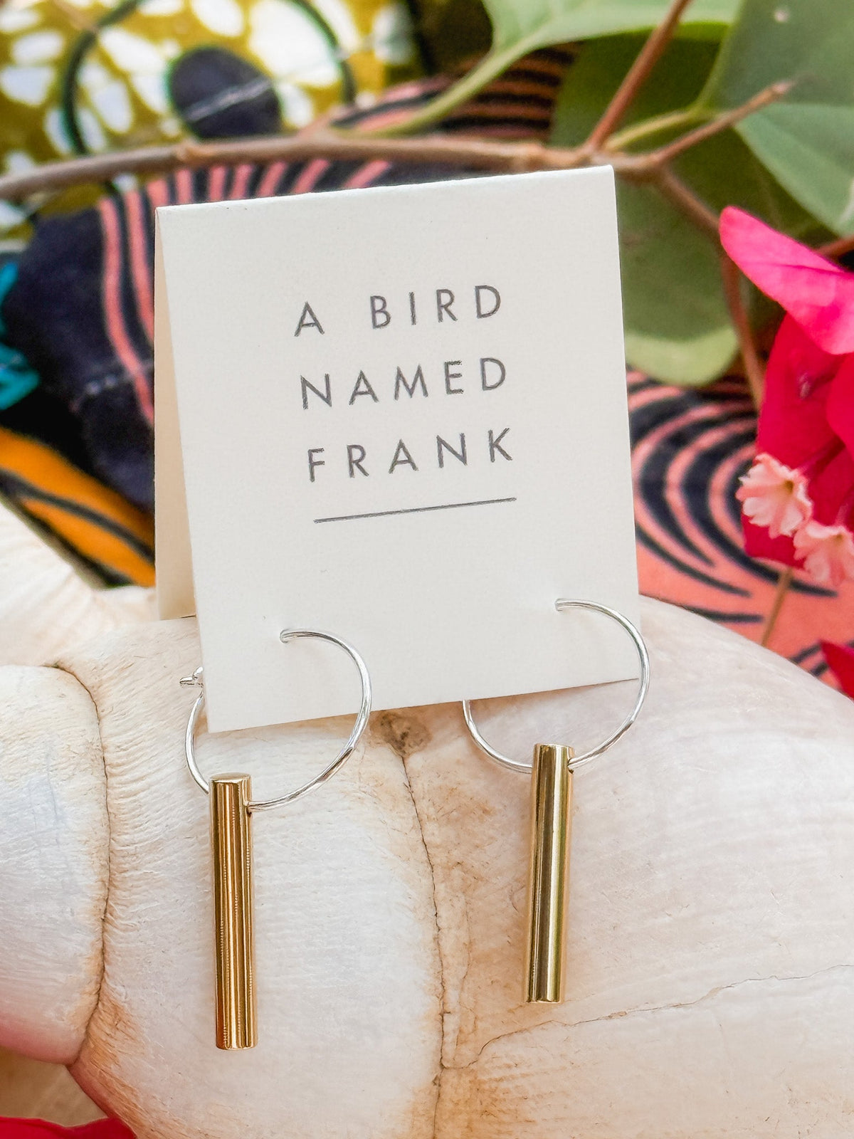 The Golden Rays Earrings are handmade hoops with gold cylindrical pendants, displayed on an A BIRD NAMED FRANK card against a vibrant background featuring leaves and flowers.