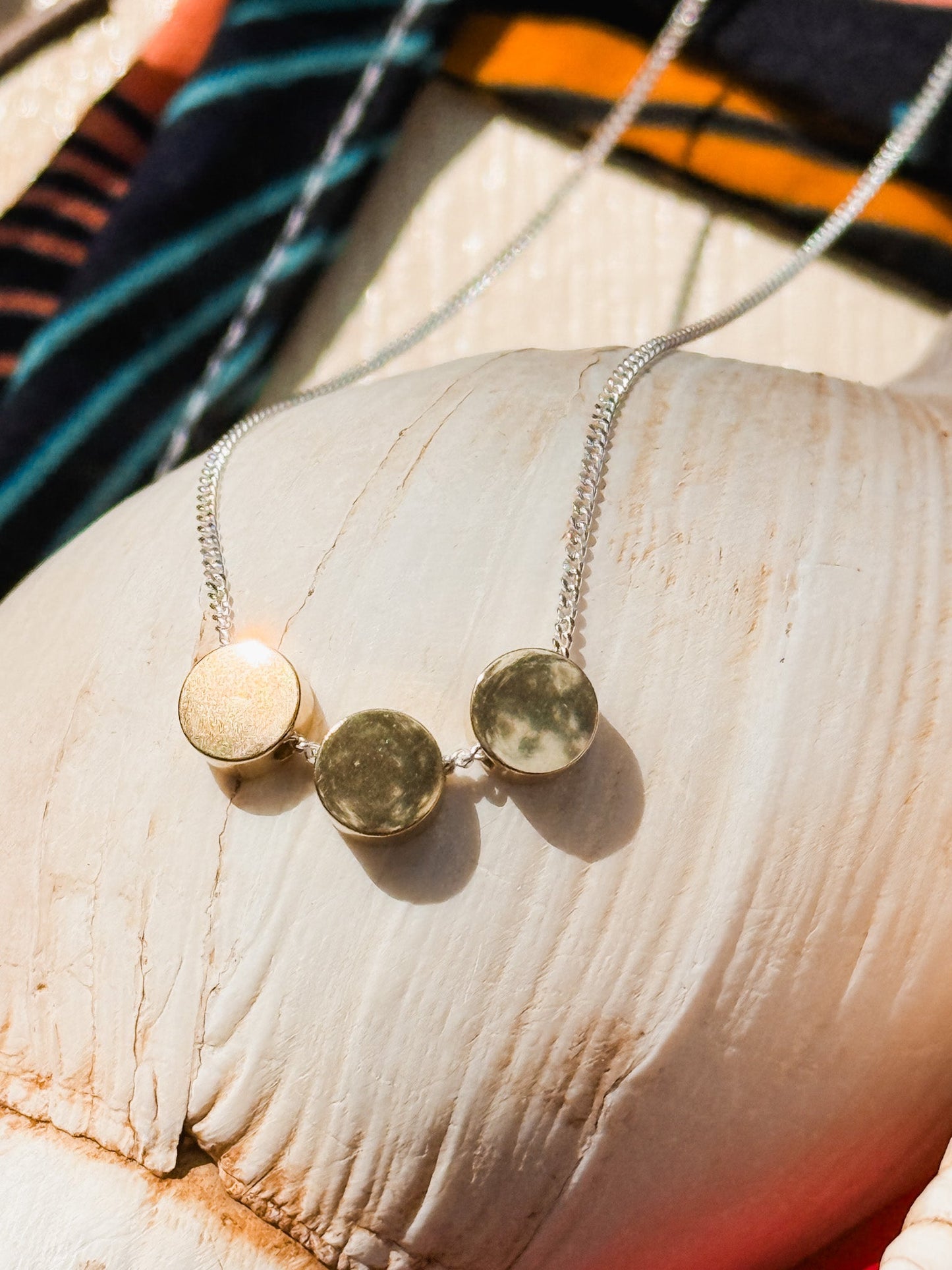The Rarara Necklace features three round moon phase pendants on a sterling silver chain, displayed on a white seashell with vibrant fabric in the background.