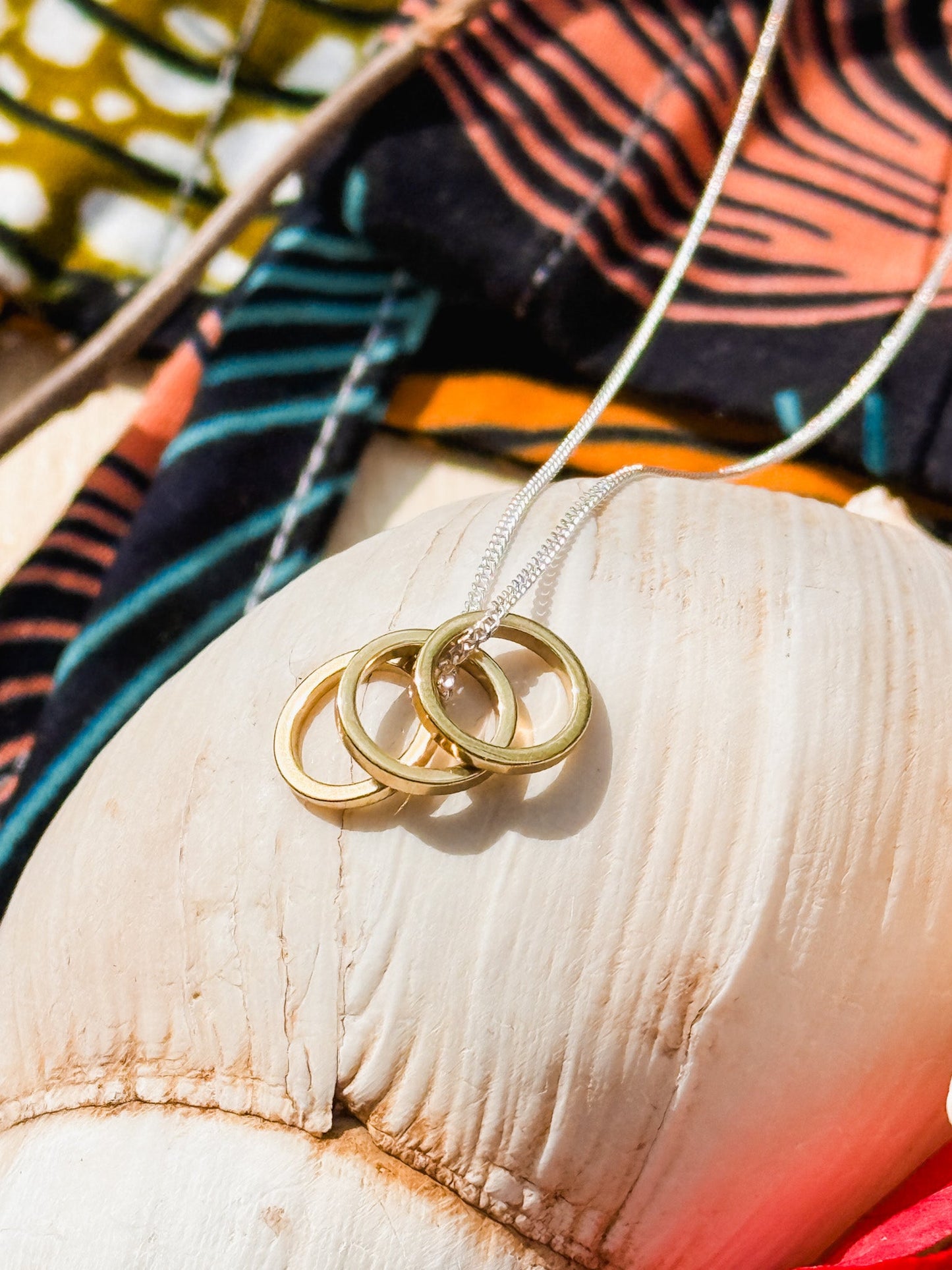 The Seeing Under Water Necklace, a minimal gold piece with three interlocking rings, rests on a large white seashell against a backdrop of colorful patterned fabric.