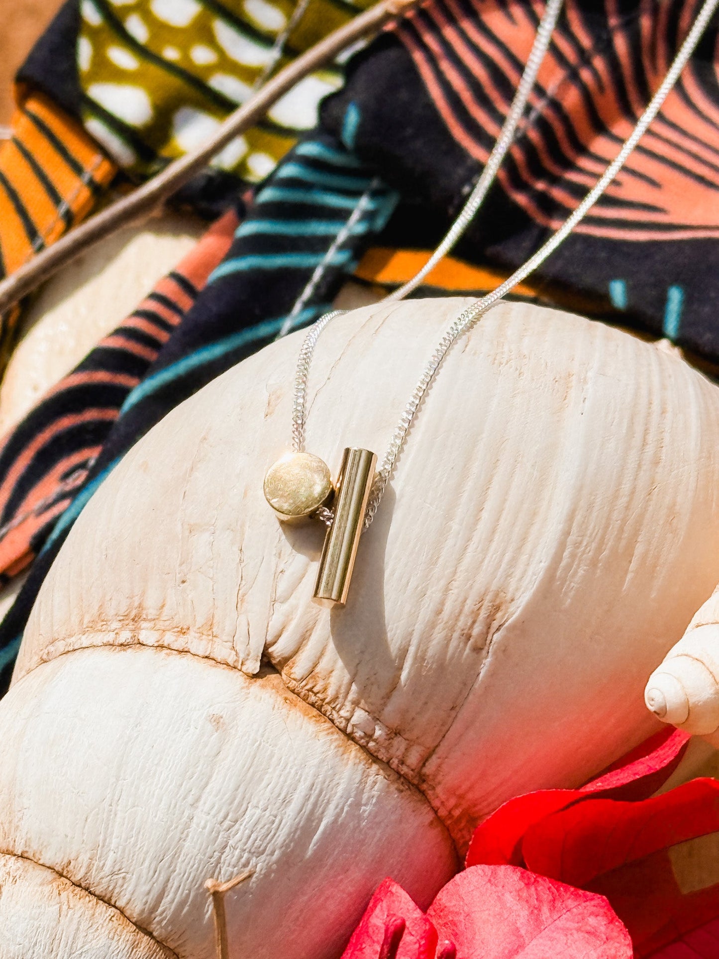 The Rock & A Hard Place Necklace, a handmade mixed metal piece with a gold cylindrical pendant and round charm on a sterling silver chain, is displayed draped over a white seashell with colorful patterned fabric in the background.