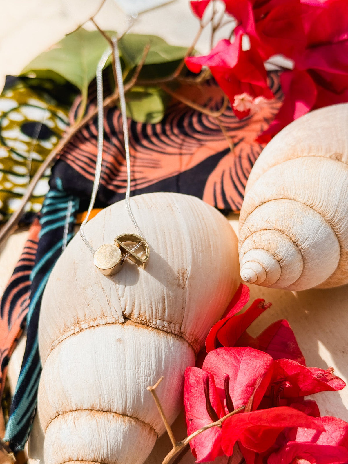 Two large white seashells rest on vibrant tropical fabric with pink flowers and green leaves. The Sun & Moon Necklace, a handmade brass and silver piece with a round pendant, sits on one shell, illuminated by sunlight in this lively South African display.