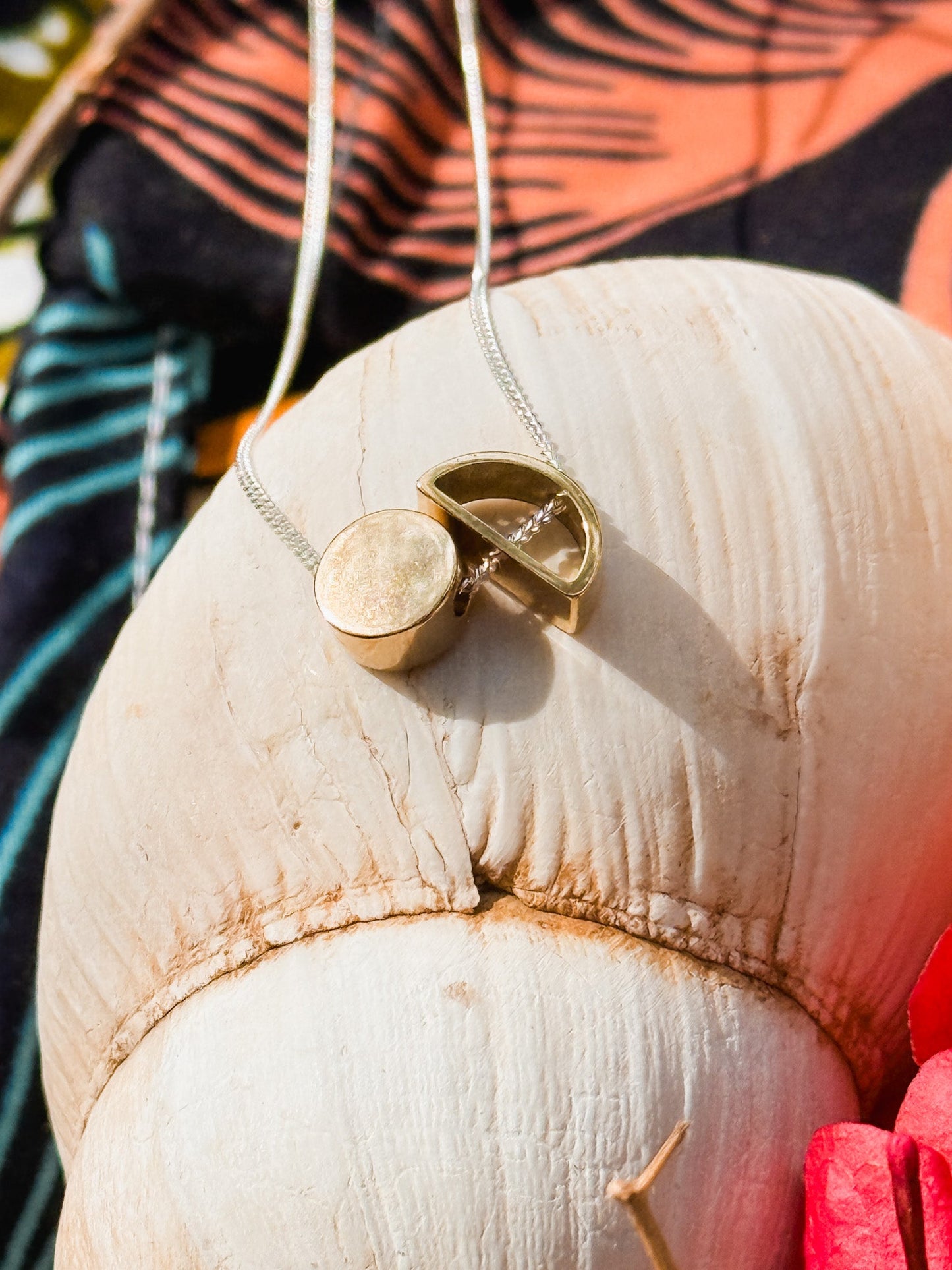 The Sun & Moon Necklace, a handmade brass and silver piece from South Africa, sits on a large white seashell. An open gold locket reveals its hollow interior, with colorful fabric and pink flowers in the background.