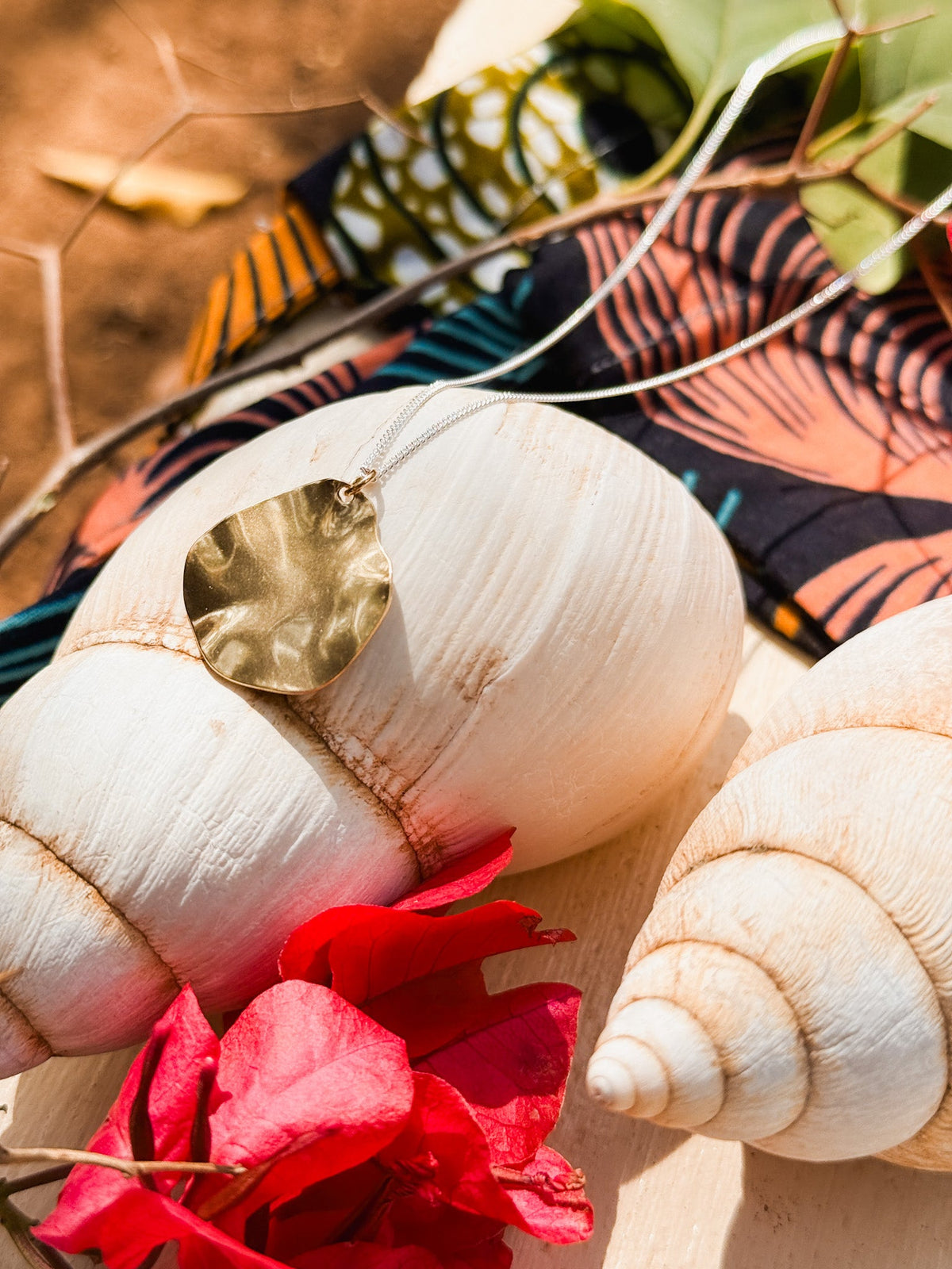 The Autumn Leaf Necklace, a handmade silver piece, rests on large white seashells with a red flower and patterned fabric in the background—highlighting Cape Town's finest jewelry, displayed on light wood.