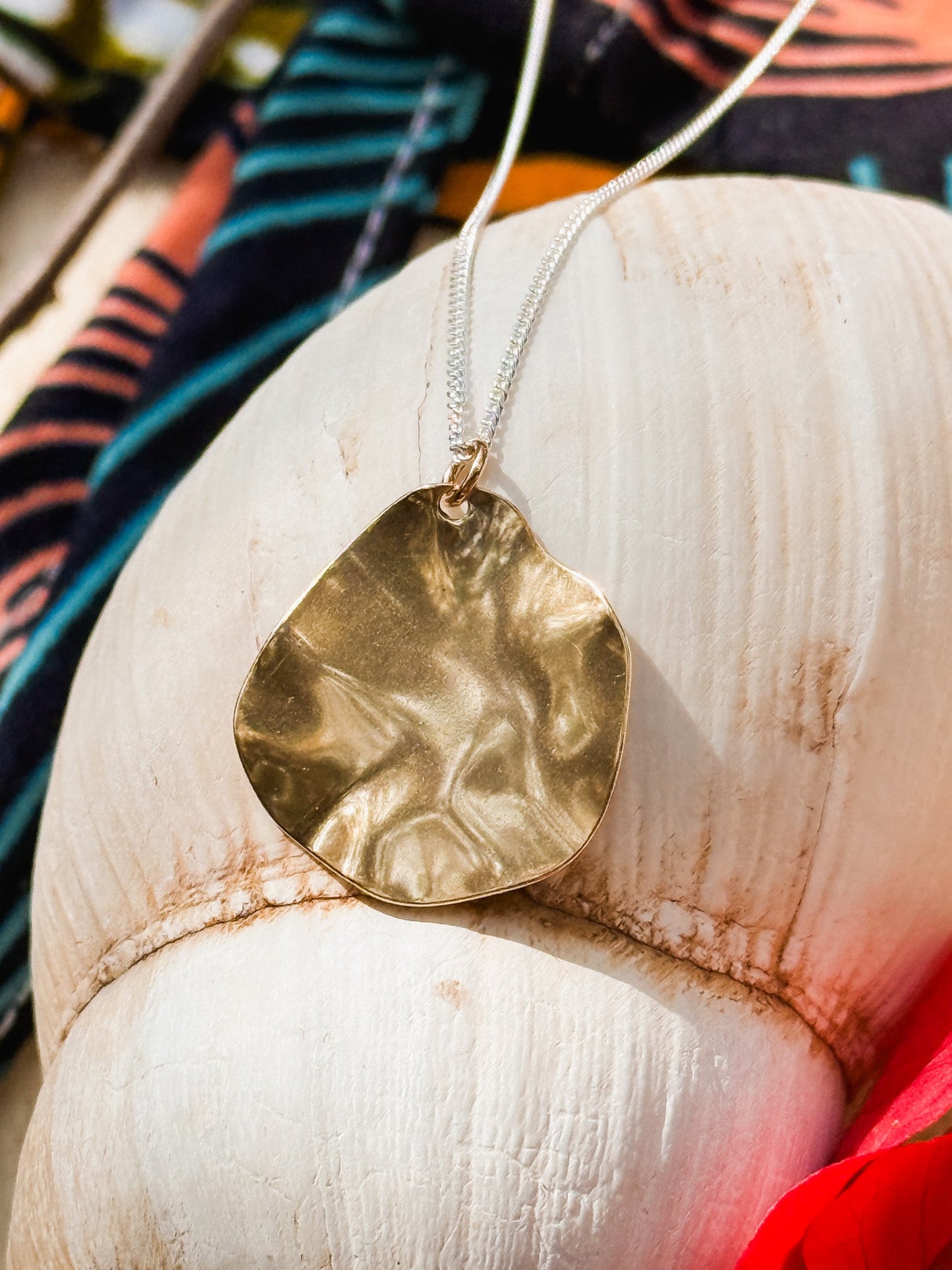 The Autumn Leaf Necklace, a gold pendant with an irregular shape, sits on a large white seashell with colorful fabric behind it, showcasing the distinctive style of Cape Town jewelry.