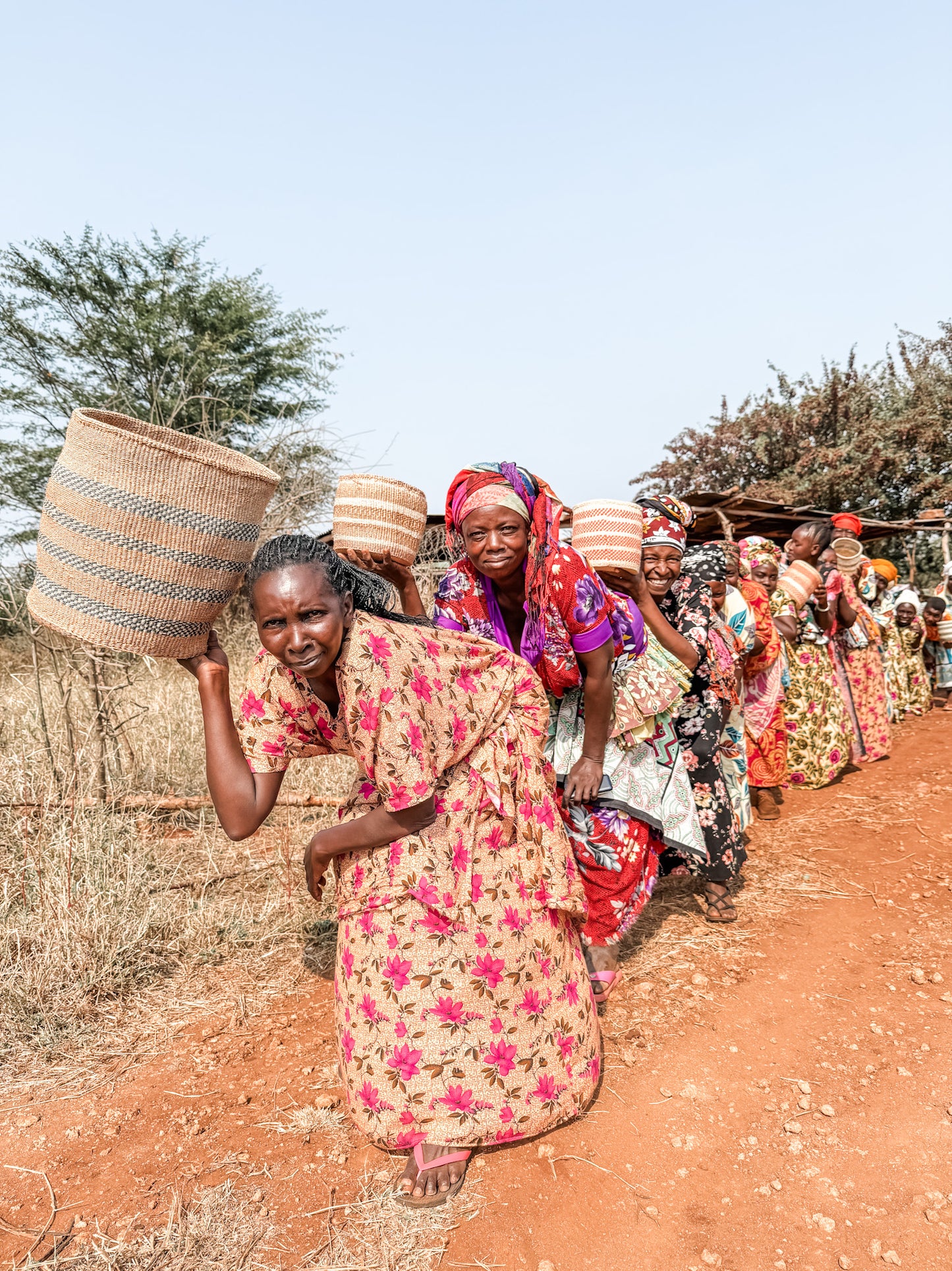 A group of women in colorful dresses and headscarves walk along a dirt path, carrying woven baskets on their backs in a rural setting with trees and dry grass—an authentic scene from the Cycling Safari experience.