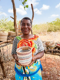A woman outdoors smiles as she holds a Thick Weave Sisal Basket. She wears a colorful dress with an orange flower print, while other handcrafted African baskets are displayed behind her on a wooden rack among trees under a partly cloudy sky.