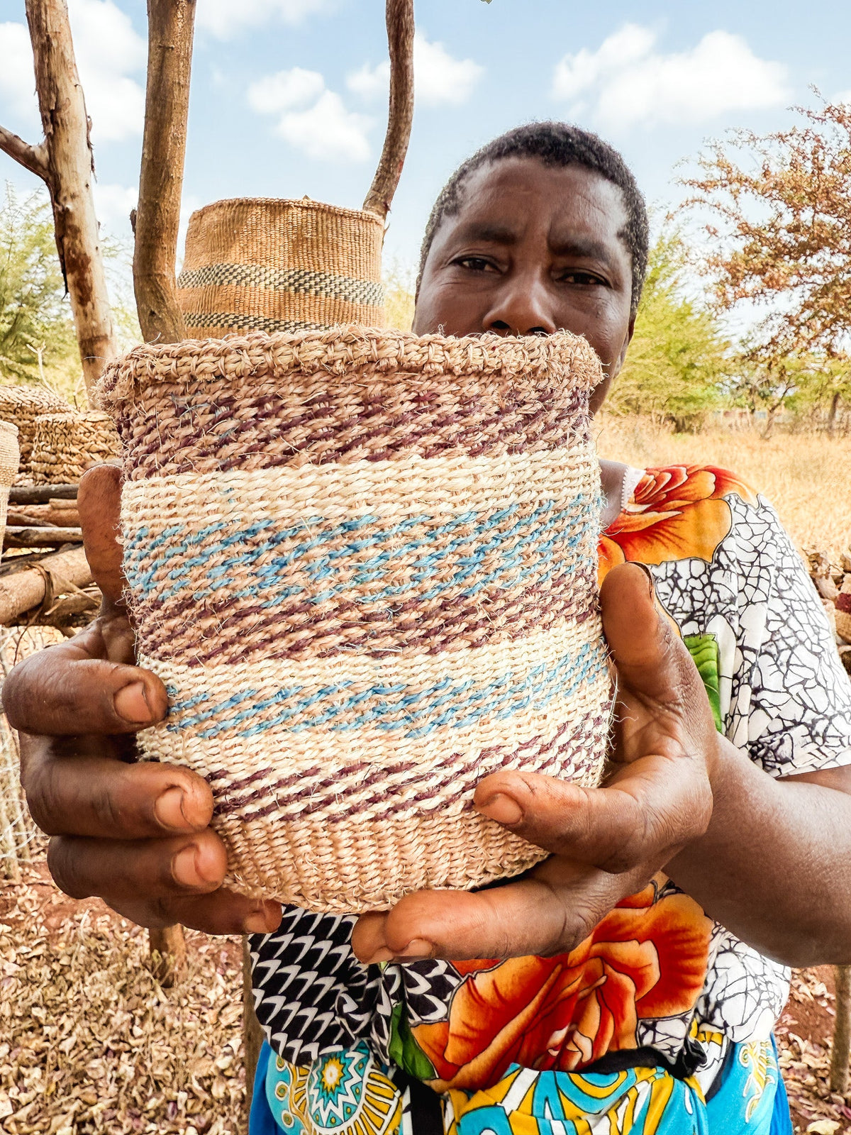 A person in colorful clothing holds up a large Thick Weave Sisal Basket with cream, brown, and light blue patterns, outdoors among trees and dry grass under a blue sky.