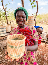 A smiling woman in a patterned dress and green knit hat displays the Thick Weave Sisal Basket toward the camera. A baby in a red cloth peeks over her shoulder, with outdoor market stalls and trees visible in the background.