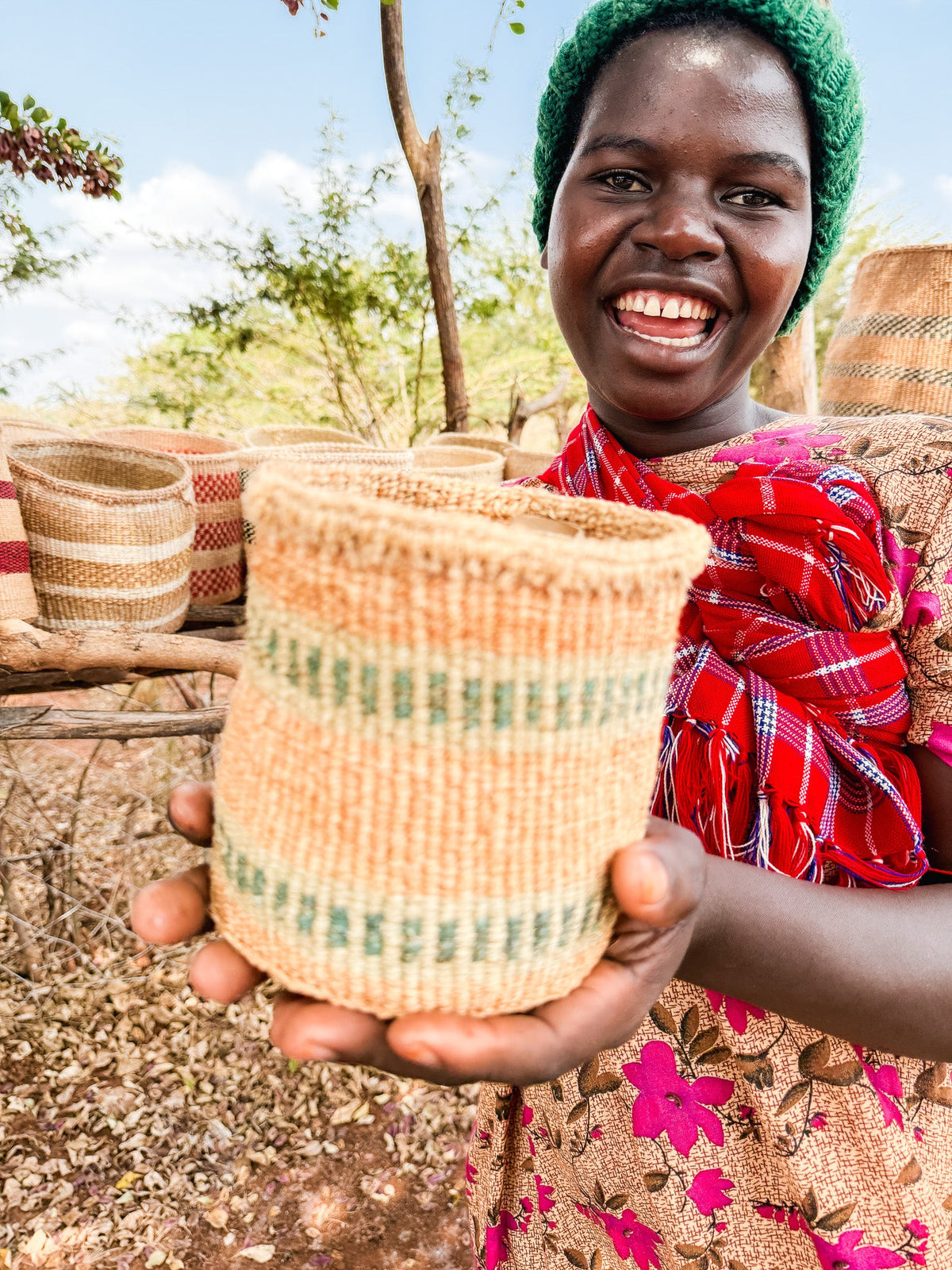 A smiling person in a green hat and red scarf holds a Thick Weave Sisal Basket toward the camera, with more fair trade baskets and trees visible in the sunny background.