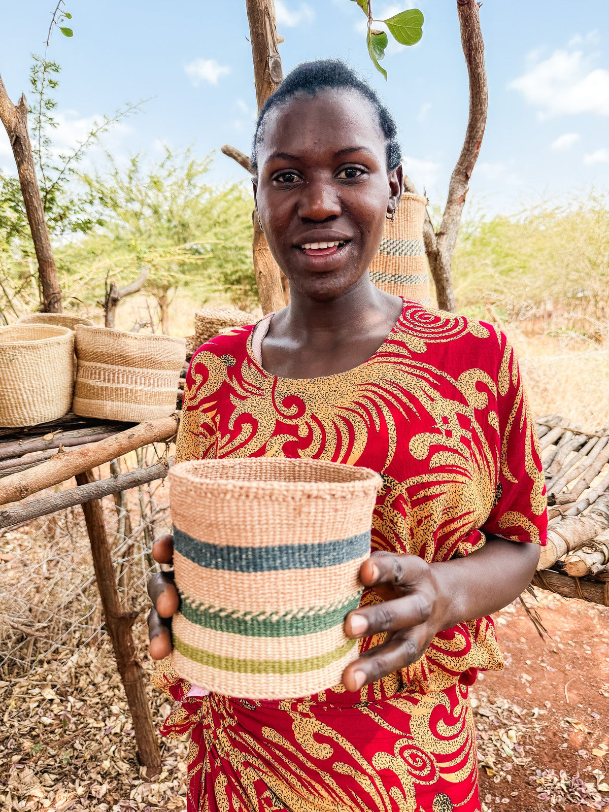 A smiling woman outdoors holds a Thick Weave Sisal Basket with green and beige stripes. More handcrafted African baskets hang on a rustic wooden stand behind her, with dry shrubs and trees in the background.
