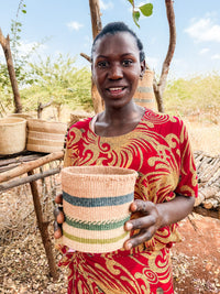 A smiling woman outdoors holds a Thick Weave Sisal Basket with green and beige stripes. More handcrafted African baskets hang on a rustic wooden stand behind her, with dry shrubs and trees in the background.