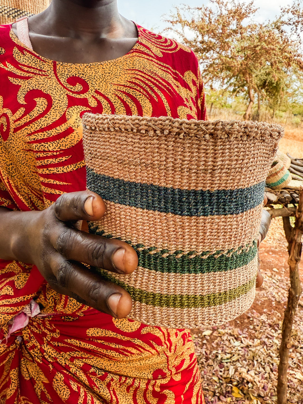 A person holds a Thick Weave Sisal Basket with blue and green stripes while wearing a vibrant red and yellow patterned garment, standing outdoors near dry grass and trees.
