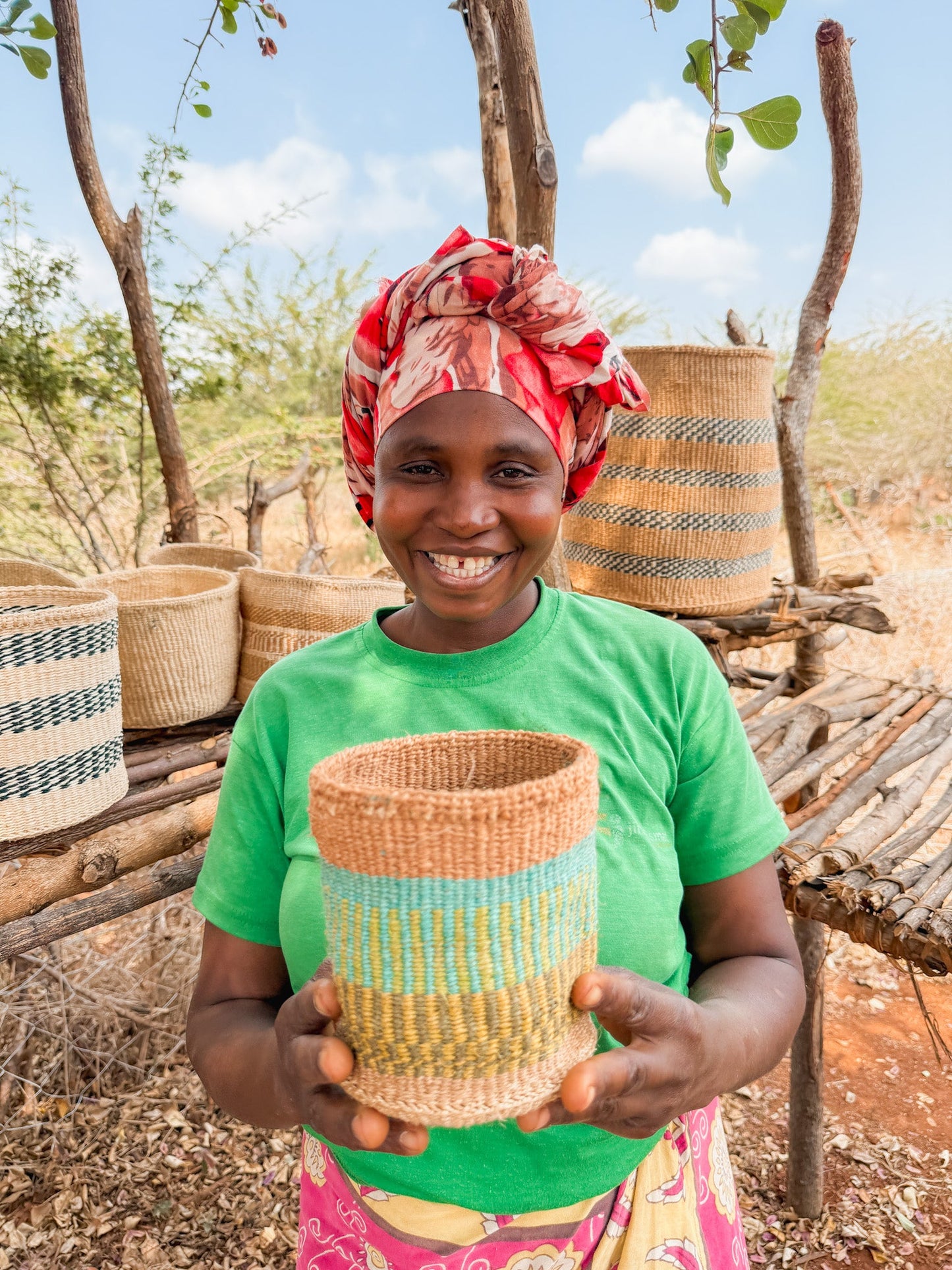 A smiling woman in a colorful headscarf and green shirt holds a Thick Weave Sisal Basket. She stands outdoors by shelves displaying fair trade baskets, with trees and a bright sky in the background.