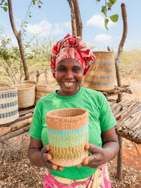 A smiling woman in a colorful headscarf and green shirt holds a Thick Weave Sisal Basket. She stands outdoors by shelves displaying fair trade baskets, with trees and a bright sky in the background.