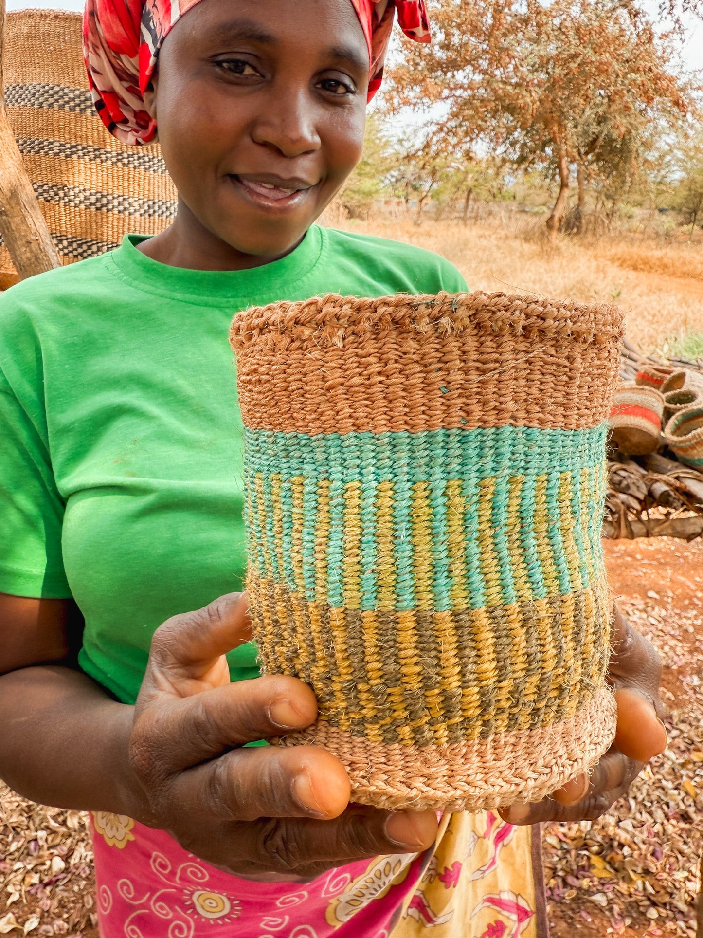 A woman in a green shirt and patterned skirt smiles outdoors as she holds up a colorful Thick Weave Sisal Basket, with more fair trade baskets and dry trees visible in the background.