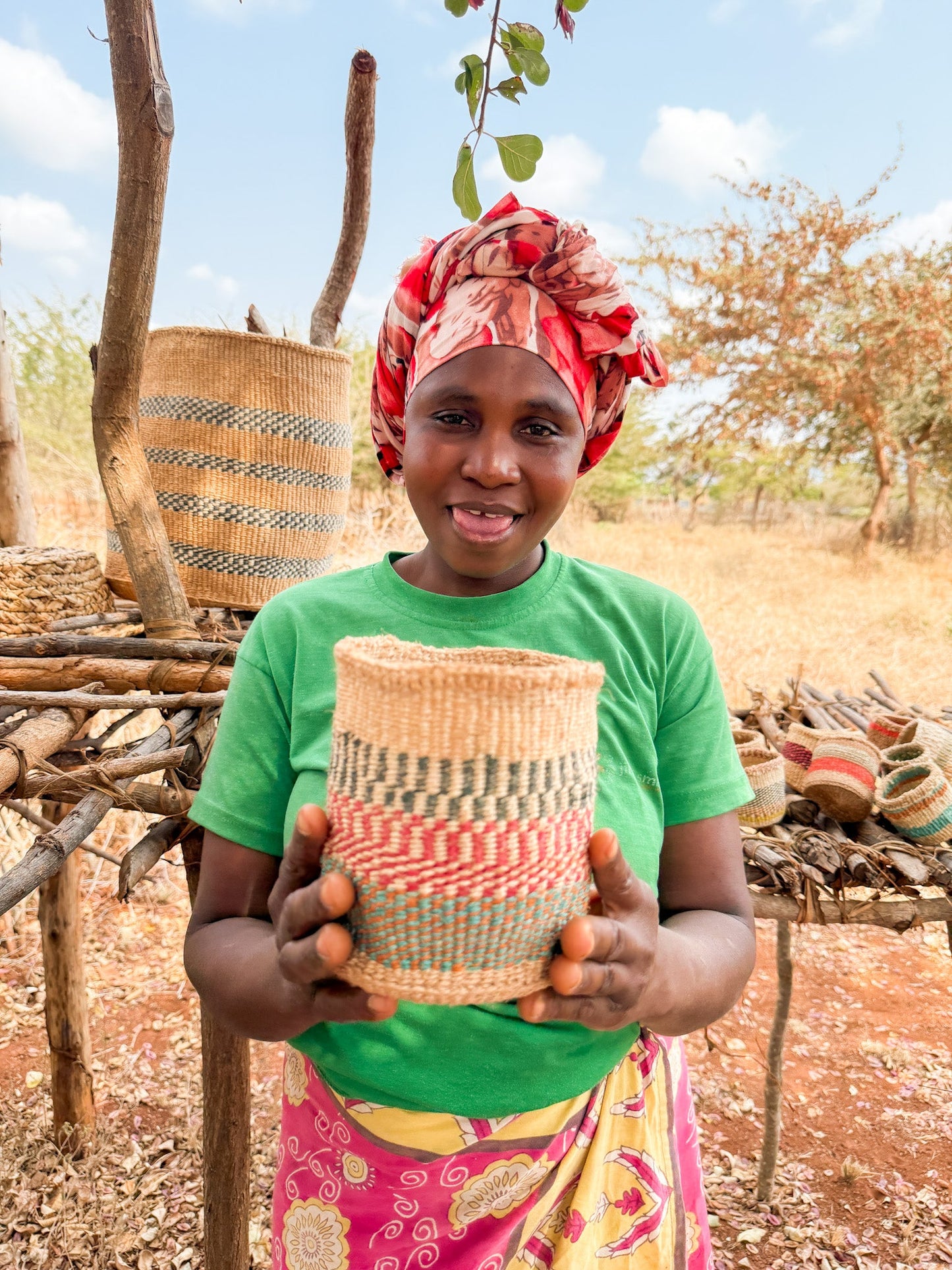 A smiling woman in a green shirt and colorful headscarf holds a Thick Weave Sisal Basket outdoors, with more handcrafted African baskets and dry trees in the background.