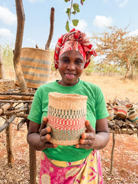 A smiling woman in a green shirt and colorful headscarf holds a Thick Weave Sisal Basket outdoors, with more handcrafted African baskets and dry trees in the background.