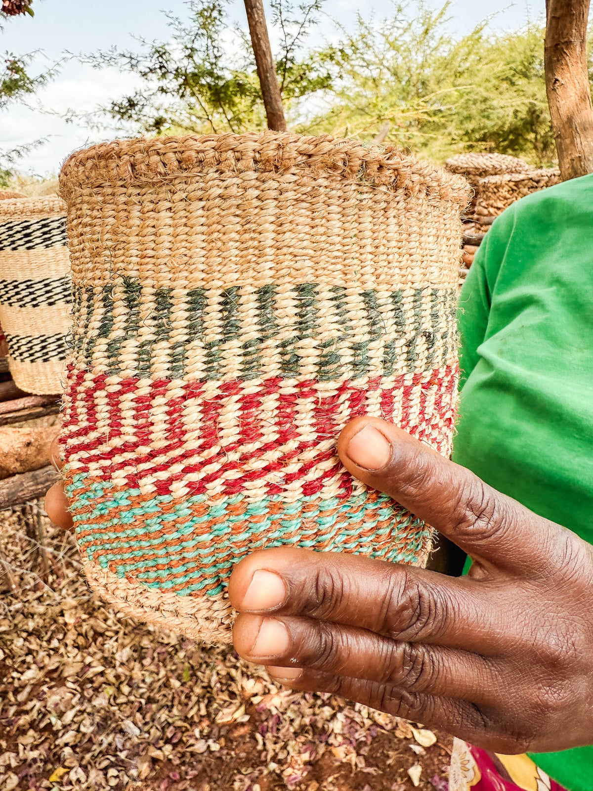 Someone holds a Thick Weave Sisal Basket, featuring vibrant natural, red, and green patterns. A green shirt sleeve is visible, with trees and more fair trade baskets displayed in the background.