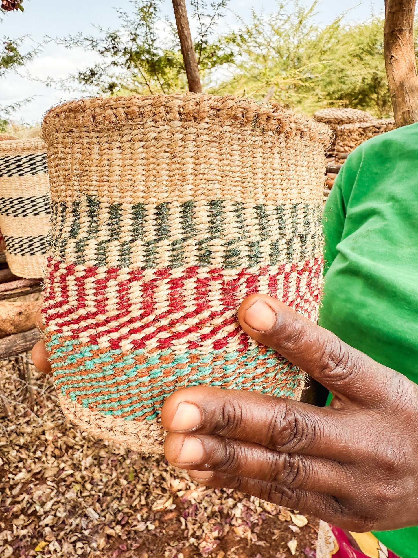 Someone holds a Thick Weave Sisal Basket, featuring vibrant natural, red, and green patterns. A green shirt sleeve is visible, with trees and more fair trade baskets displayed in the background.