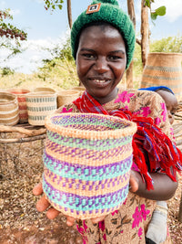 A smiling person in a green hat and patterned clothes holds a Thick Weave Sisal Basket toward the camera, with more fair trade baskets displayed on a wooden table outdoors.