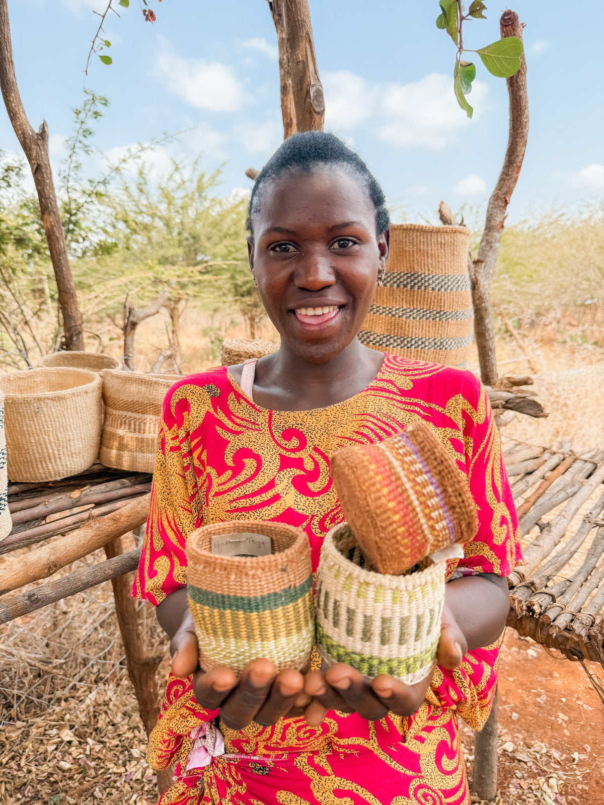 A smiling woman in a vibrant red and yellow dress holds out three Thick Weave Sisal Baskets. Behind her, more handcrafted African baskets are displayed on a wooden stand outdoors amid dry grass and trees.