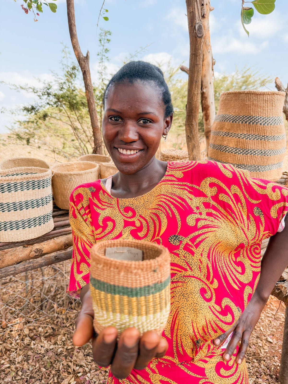 A woman in a red and yellow dress smiles while holding a Thick Weave Sisal Basket. Several handcrafted baskets are displayed on wooden shelves outdoors, with dry grass and trees in the background.