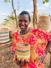 A woman in a red and yellow dress smiles while holding a Thick Weave Sisal Basket. Several handcrafted baskets are displayed on wooden shelves outdoors, with dry grass and trees in the background.