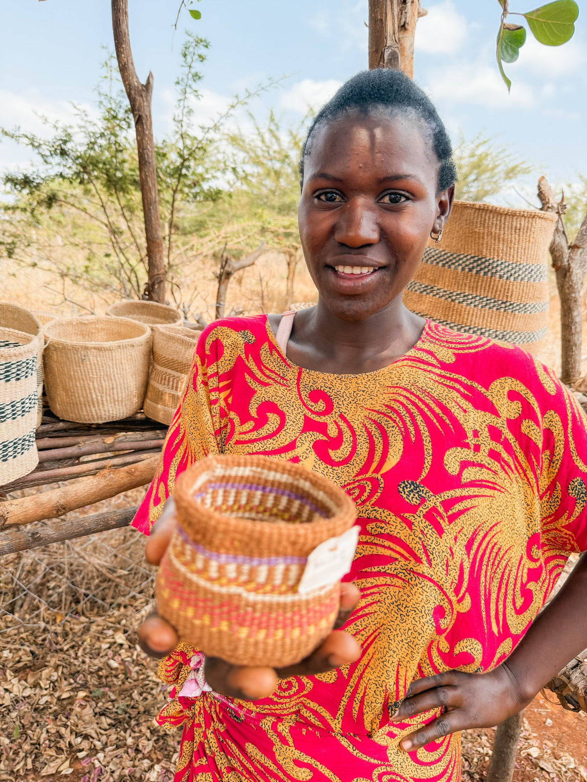 A woman in a vibrant red and yellow dress smiles as she presents a Thick Weave Sisal Basket to the camera, with more baskets arranged on a wooden stand behind her outdoors.