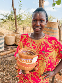 A woman in a vibrant red and yellow dress smiles as she presents a Thick Weave Sisal Basket to the camera, with more baskets arranged on a wooden stand behind her outdoors.