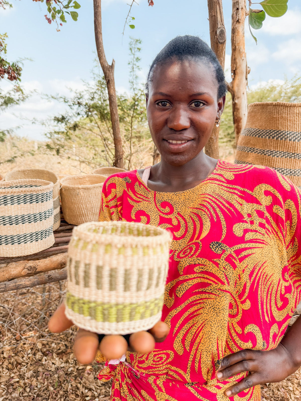 A woman in a vibrant dress presents a Thick Weave Sisal Basket toward the camera, with additional fair trade Kenyan baskets displayed on a wooden shelf behind her outdoors.
