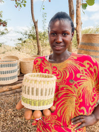 A woman in a vibrant dress presents a Thick Weave Sisal Basket toward the camera, with additional fair trade Kenyan baskets displayed on a wooden shelf behind her outdoors.