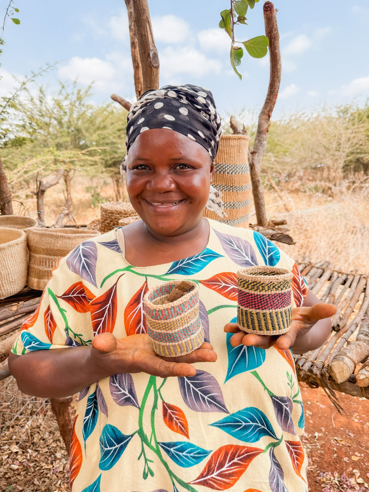 A smiling woman in a patterned dress and headscarf holds two Thick Weave Sisal Baskets. More baskets are displayed on a wooden structure behind her, outdoors in a dry, rural setting.