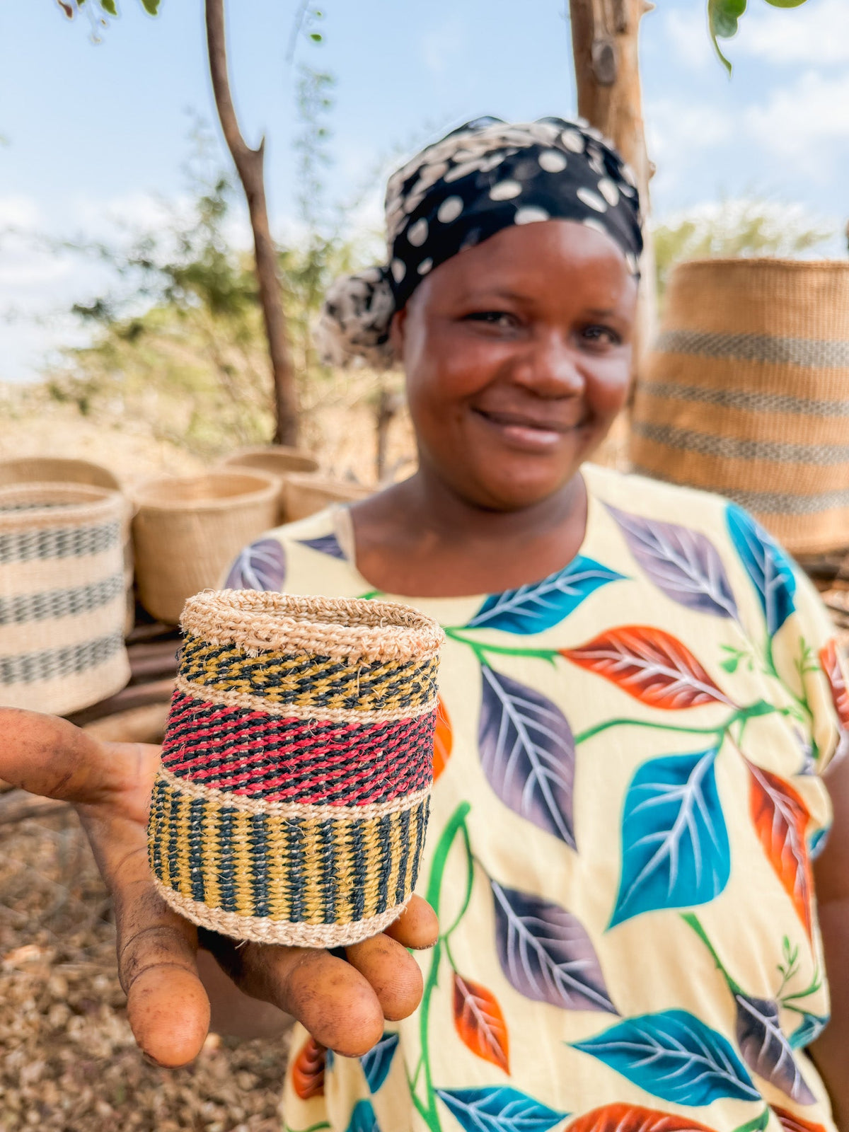 A smiling woman in a colorful dress and headscarf holds a small, handcrafted Thick Weave Sisal Basket featuring red, green, and yellow designs. Larger baskets are displayed in the background outdoors.
