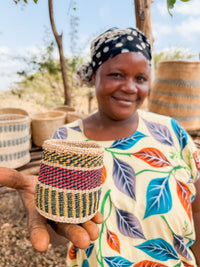 A smiling woman in a colorful dress and headscarf holds a small, handcrafted Thick Weave Sisal Basket featuring red, green, and yellow designs. Larger baskets are displayed in the background outdoors.