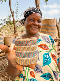 A smiling woman in a patterned dress and headscarf holds out a small, colorful Thick Weave Sisal Basket toward the camera, with larger handcrafted African baskets arranged behind her outdoors under the trees and sky.