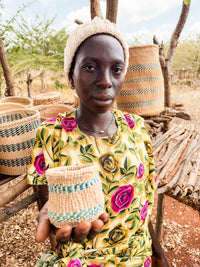 A woman in a yellow dress with pink flowers and a knit hat presents a small handcrafted Thick Weave Sisal Basket to the camera, with larger sisal baskets displayed outdoors in a rural setting behind her.