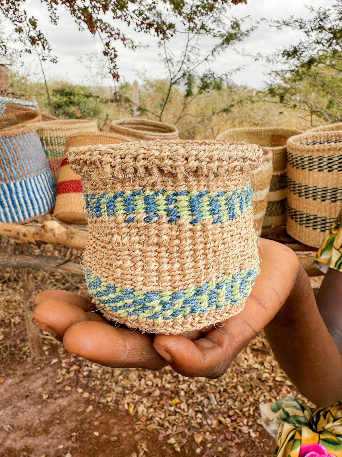 A hand holds a Thick Weave Sisal Basket with blue and green patterns. In the background, several handcrafted African baskets with unique designs are displayed outdoors among dry plants and trees.