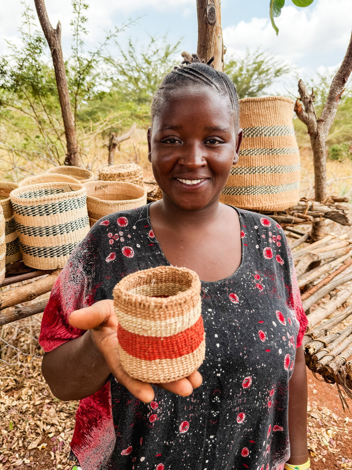A smiling woman stands outdoors, holding the Thick Weave Sisal Basket featuring red and natural stripes. Behind her, more handcrafted baskets are displayed on a wooden rack, with dry grass and scattered trees in the background.