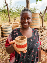 A smiling woman stands outdoors, holding the Thick Weave Sisal Basket featuring red and natural stripes. Behind her, more handcrafted baskets are displayed on a wooden rack, with dry grass and scattered trees in the background.