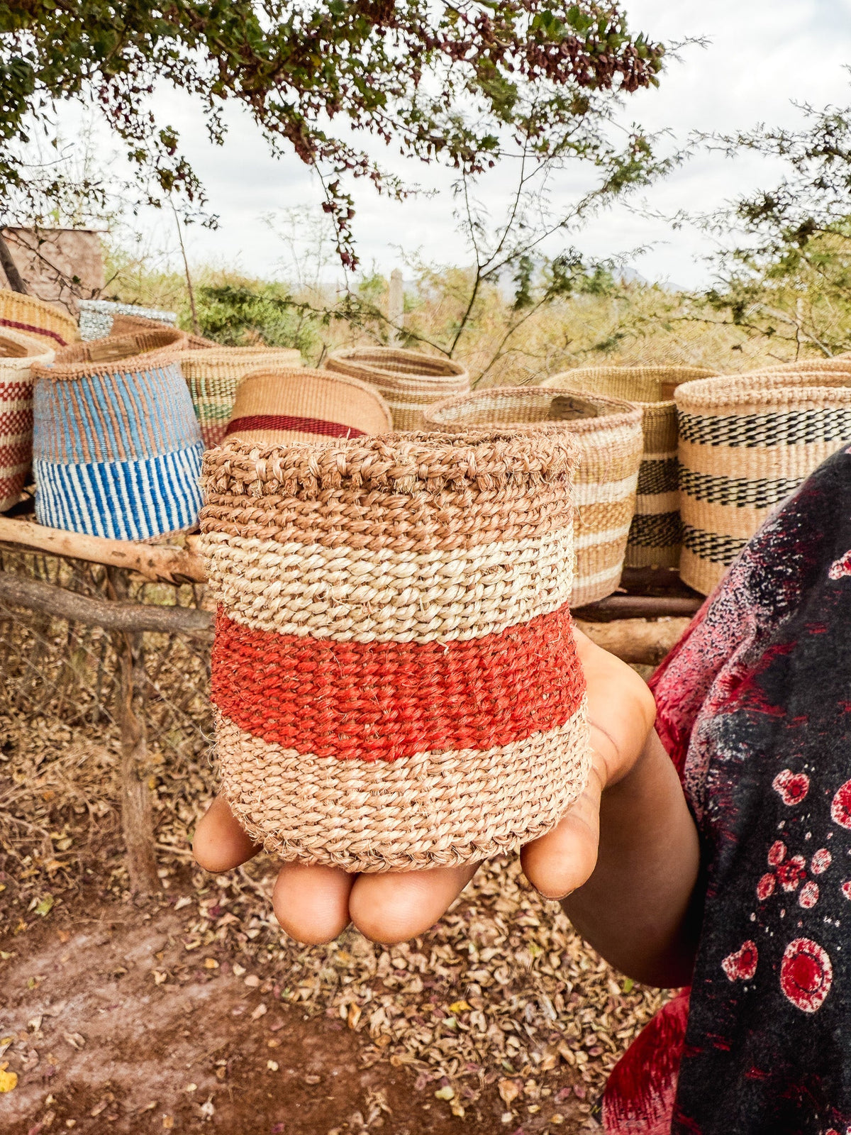 A person holds a Thick Weave Sisal Basket with a bold red stripe, while more vibrant handcrafted baskets are displayed on a wooden rack outdoors among trees and dry leaves.