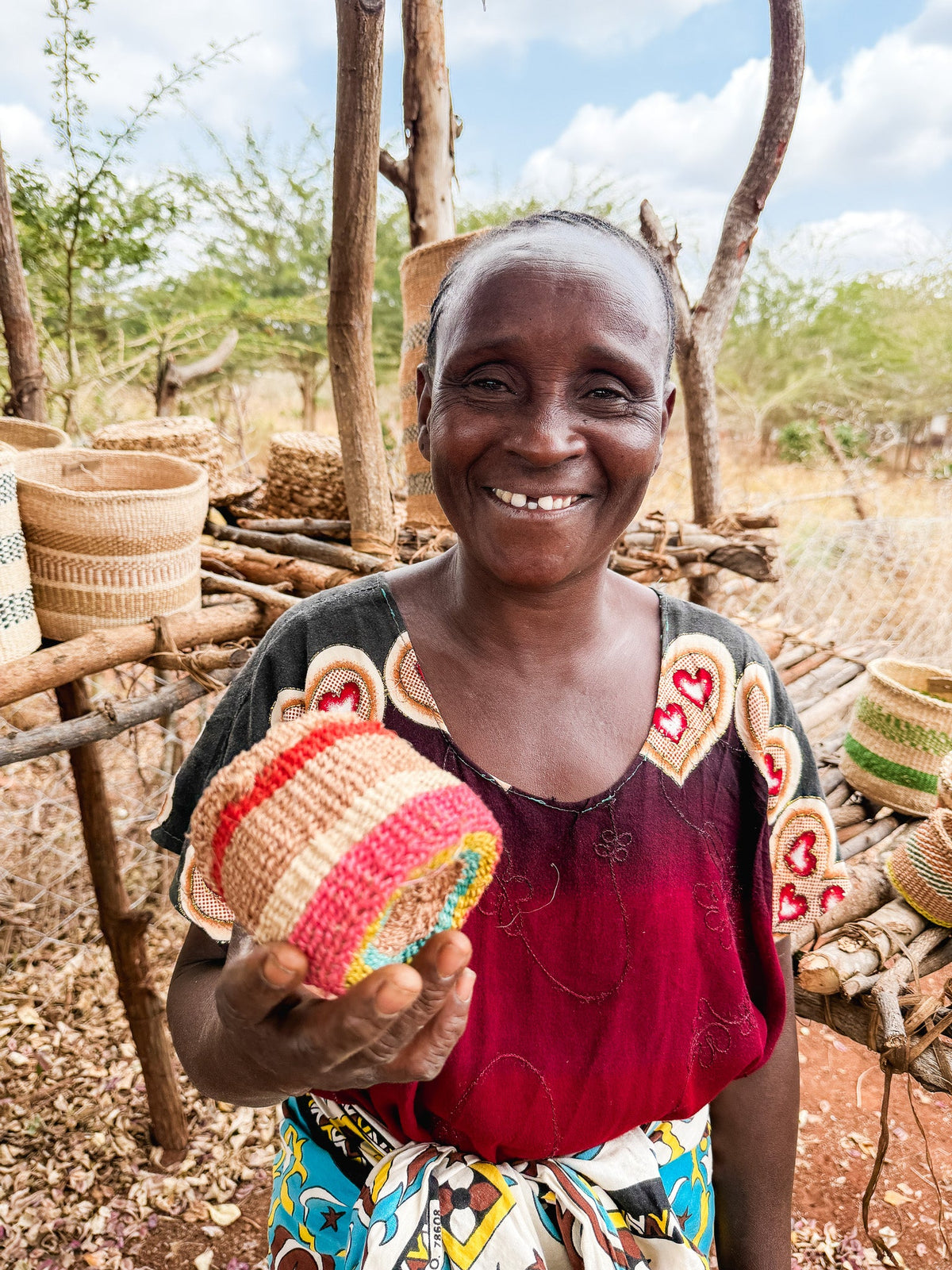 A smiling woman stands outdoors holding a Thick Weave Sisal Basket. Dressed in a red heart-patterned top, she is beside a display of handcrafted African baskets made from natural materials, with trees and a fence behind her.