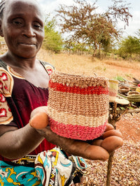 A smiling woman outdoors holds out the Thick Weave Sisal Basket, featuring bold red and natural stripes, with dry grass and trees in the background.