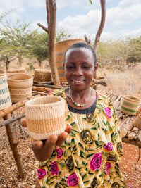 A woman in a floral dress smiles while holding a Thick Weave Sisal Basket toward the camera. Several other handcrafted African baskets are arranged on a wooden stand outdoors, with trees and dry grass in the background.