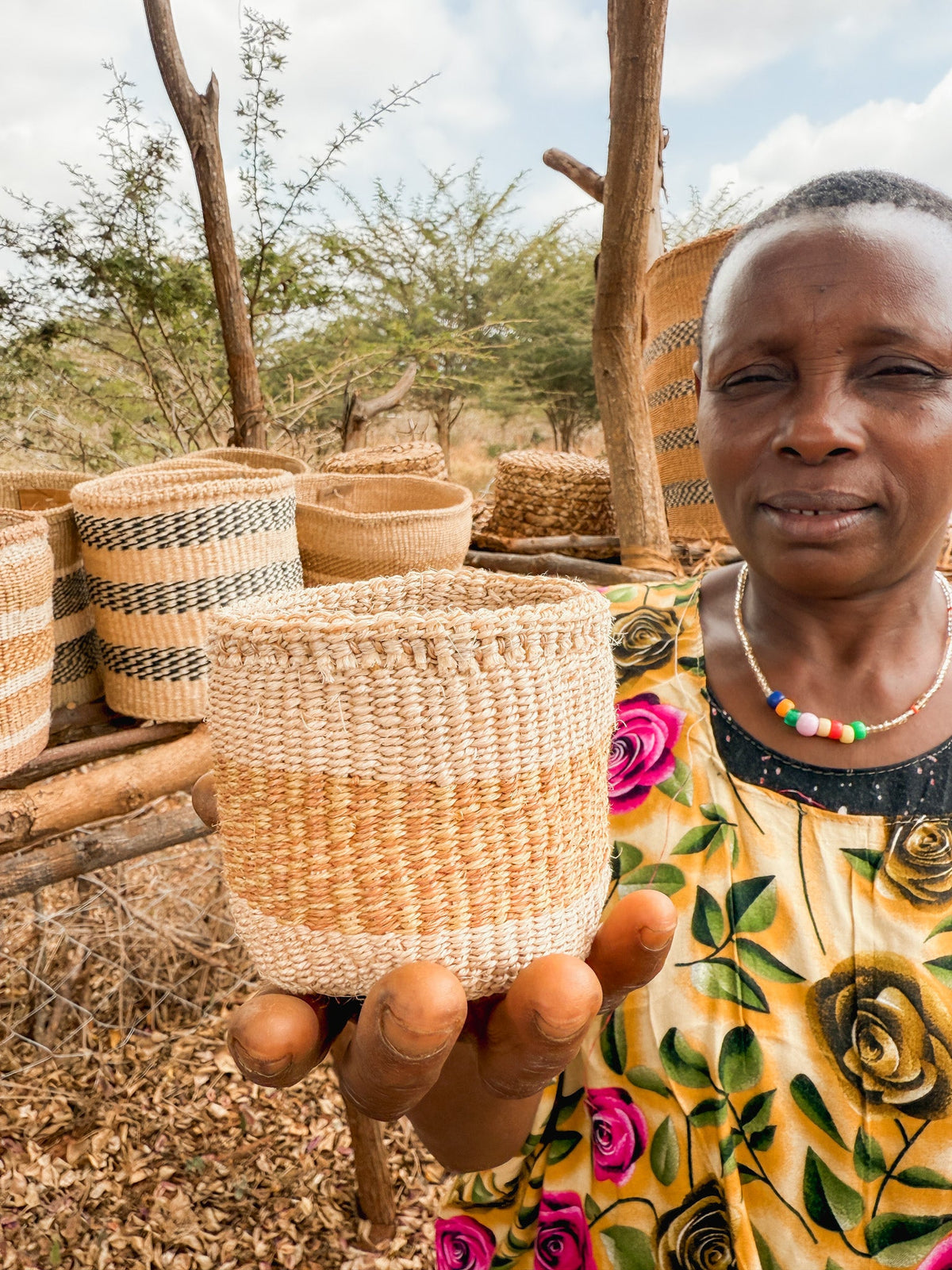 A woman in a vibrant floral shirt and beaded necklace holds a handcrafted Thick Weave Sisal Basket toward the camera, with several fair trade Kenyan baskets displayed on wooden shelves behind her in an outdoor setting.