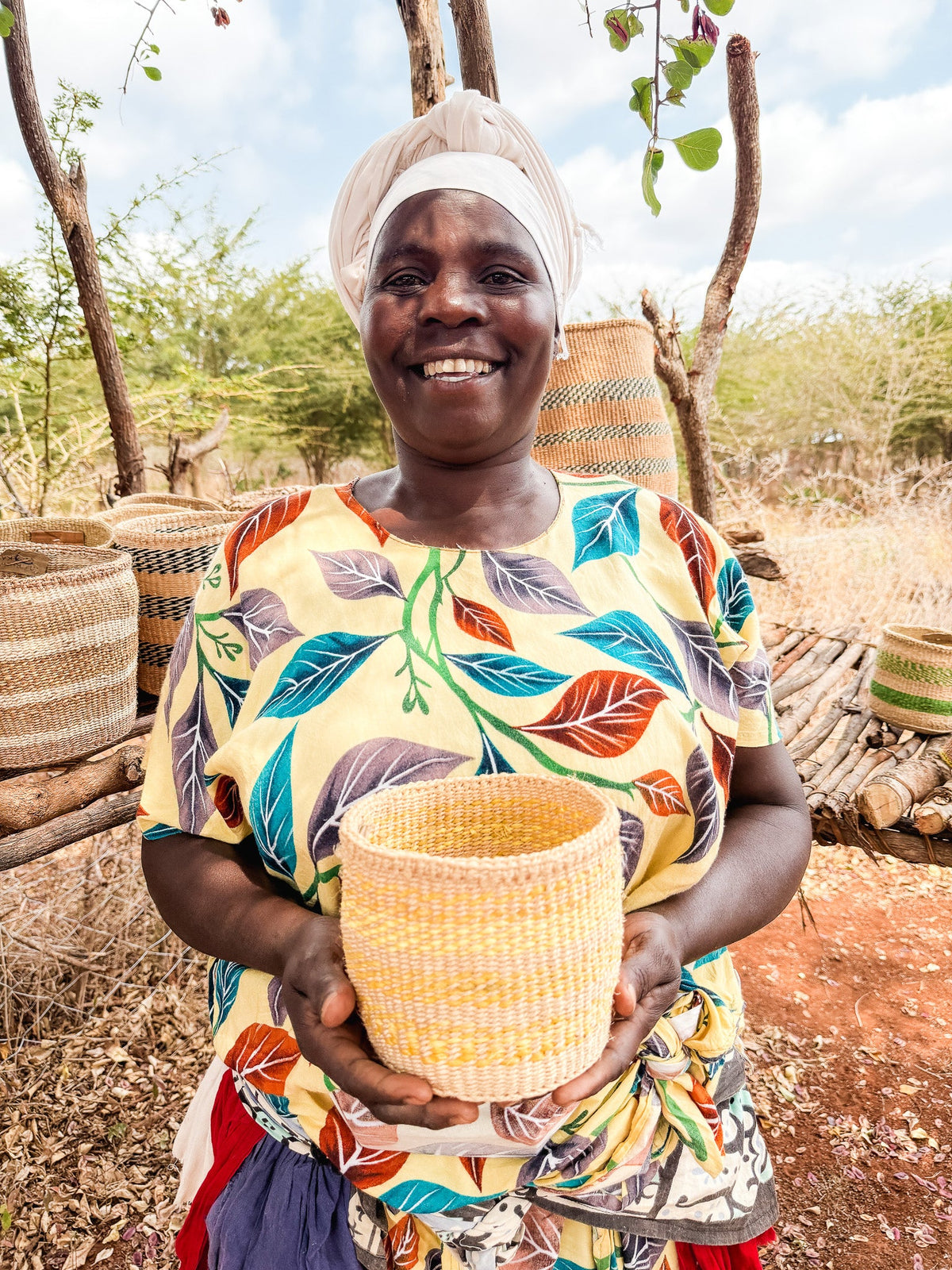 A smiling woman in a colorful leaf-patterned dress and white headscarf holds a Thick Weave Sisal Basket outdoors, with more baskets displayed on wooden stands and dry trees in the background.
