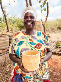 A smiling woman in a colorful leaf-patterned dress and white headscarf holds a Thick Weave Sisal Basket outdoors, with more baskets displayed on wooden stands and dry trees in the background.