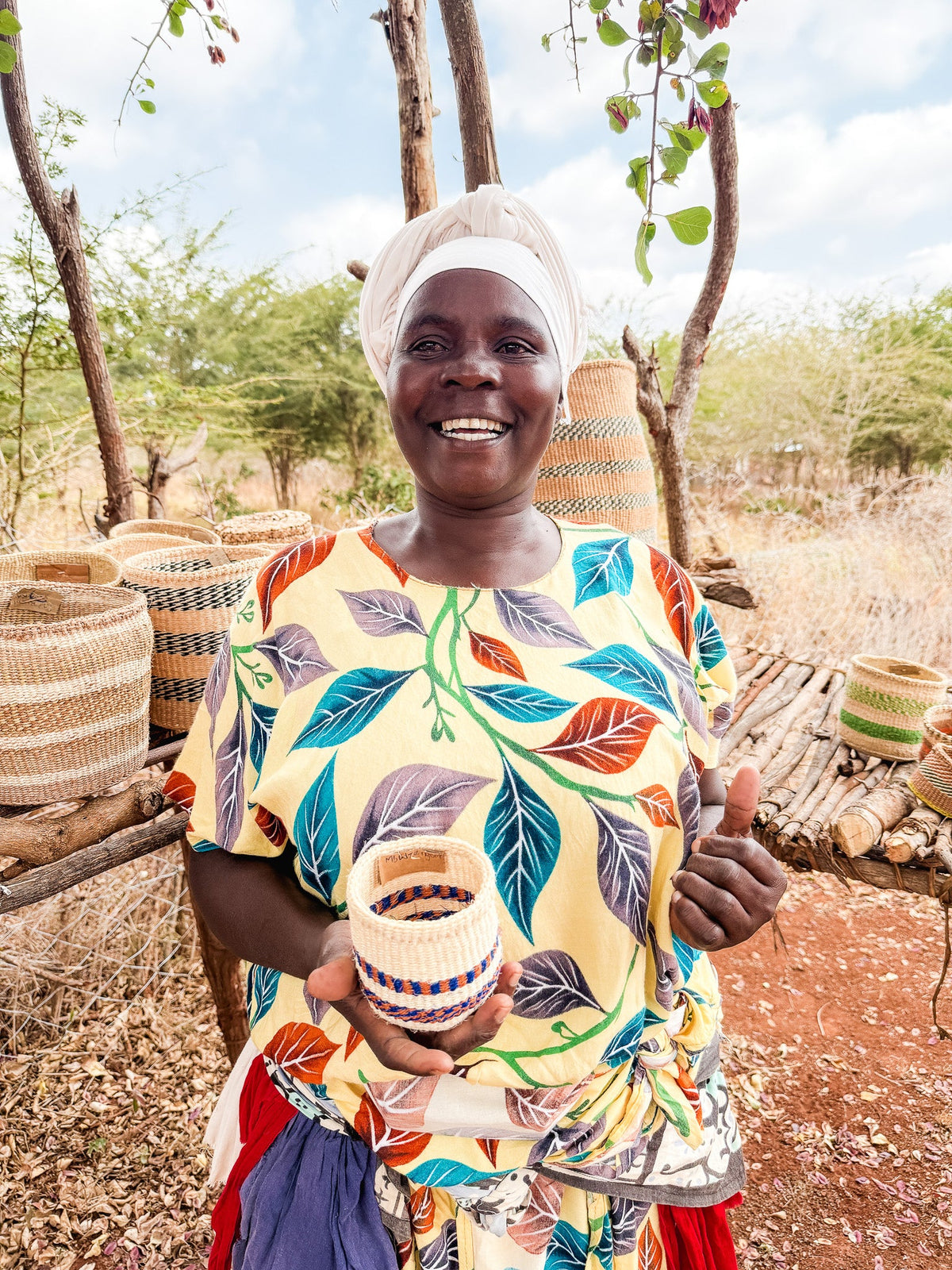 A smiling woman in a colorful leaf-patterned dress and white headwrap stands outdoors, holding a Thick Weave Sisal Basket. Fair trade baskets are arranged on wooden shelves behind her.