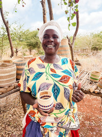 A smiling woman in a colorful leaf-patterned dress and white headwrap stands outdoors, holding a Thick Weave Sisal Basket. Fair trade baskets are arranged on wooden shelves behind her.